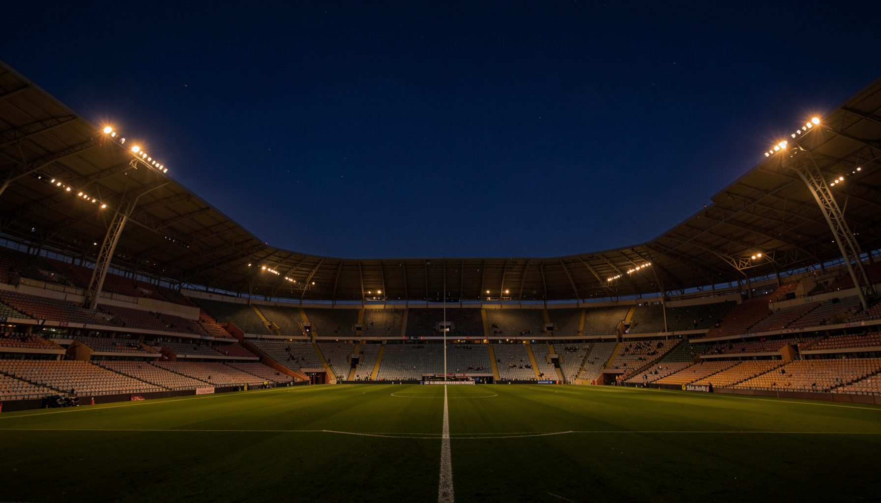 Atmospheric wide shot of an empty sports stadium at dusk with deep navy sky and warm amber field lights glowing softly